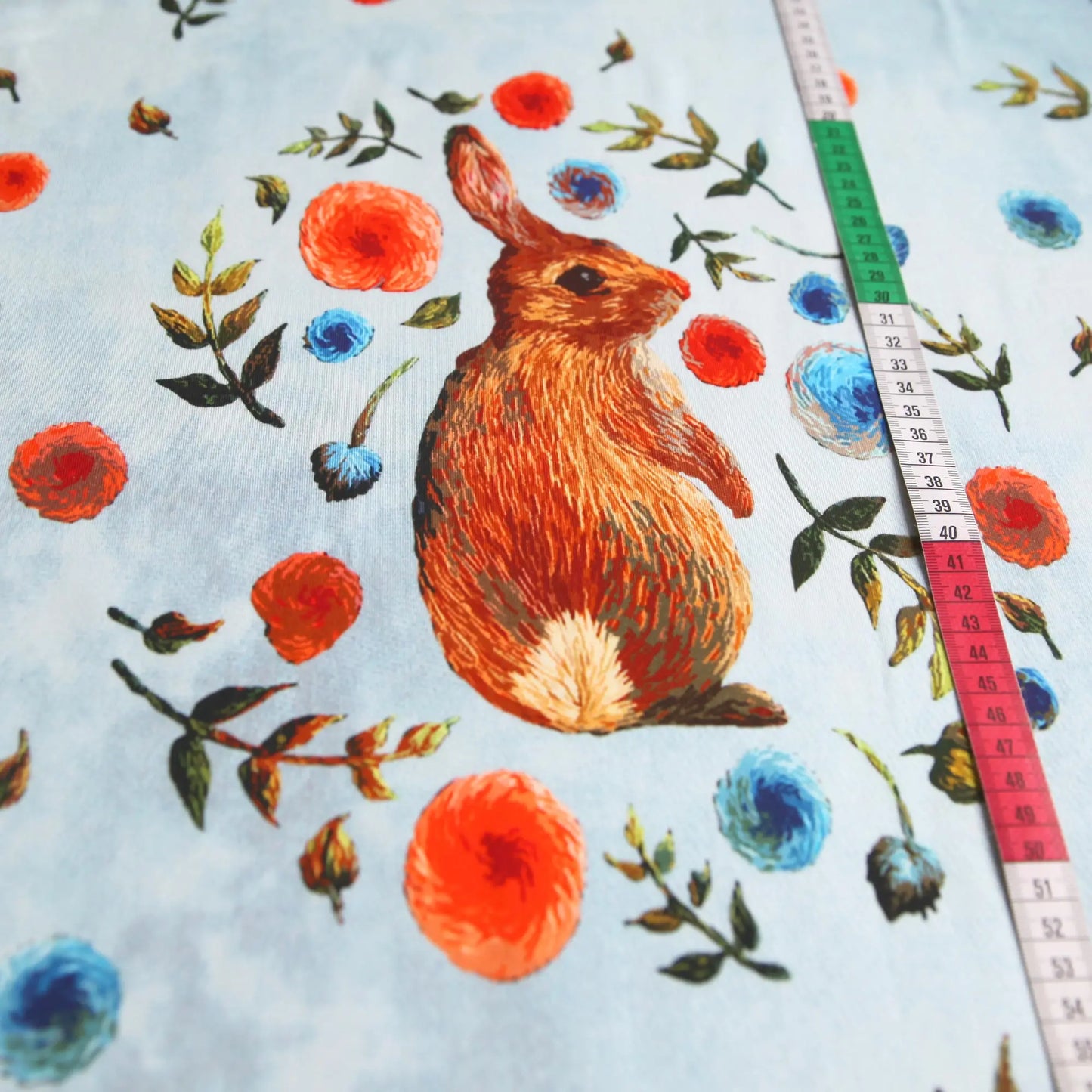 Hellblauer Baumwolljersey mit braunem Kaninchen und bunten Blumenmotiven, Metermaß an der Seite.