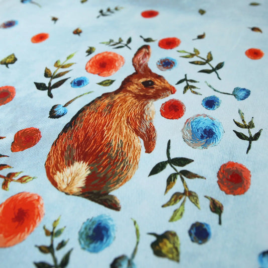 Hellblauer Baumwolljersey mit braunem Kaninchen und bunten Blumenmotiven.