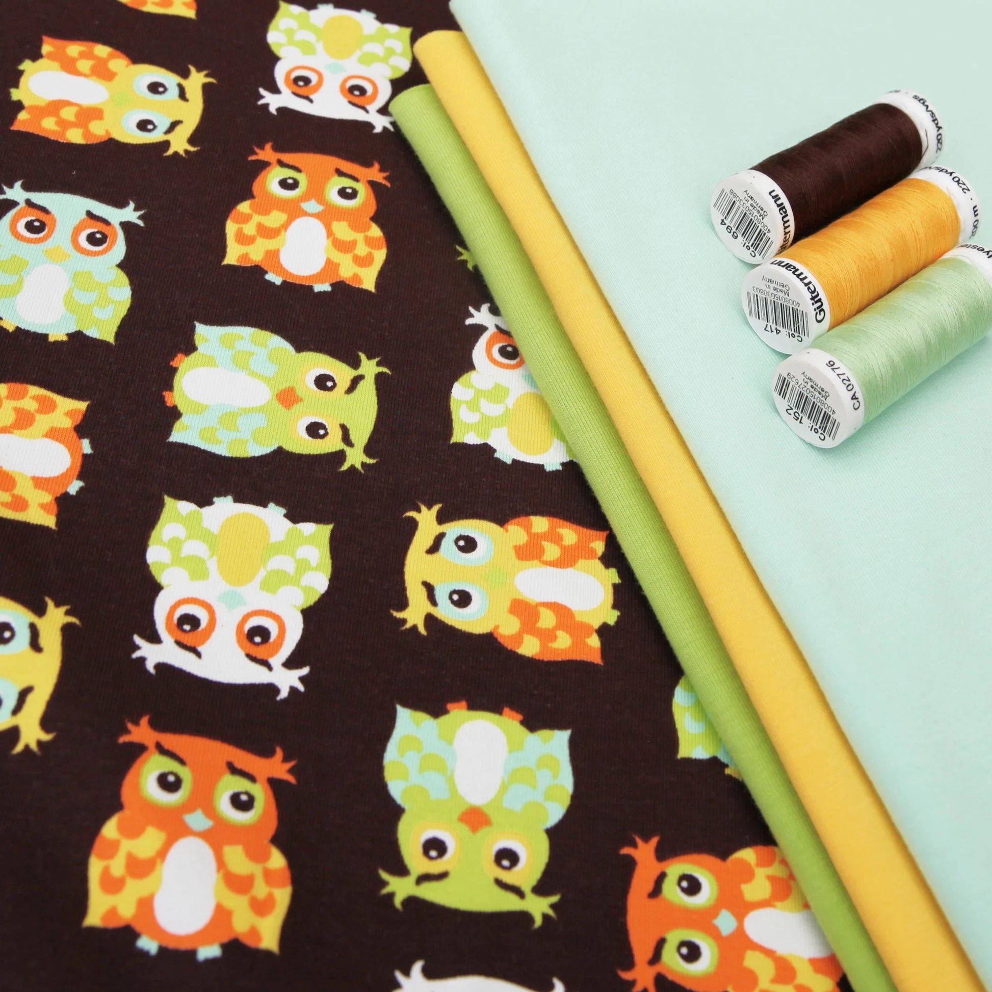 Baumwolljersey mit bunten Eulen auf braunem Hintergrund, daneben gelber, grüner und mintfarbener Stoff sowie passende.